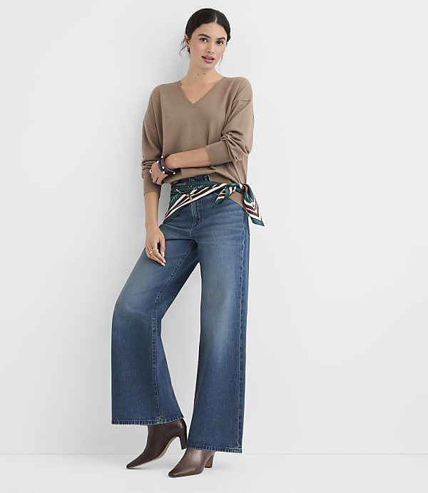 A woman in a brown v-neck sweater and wide-leg jeans stands against a plain white background.