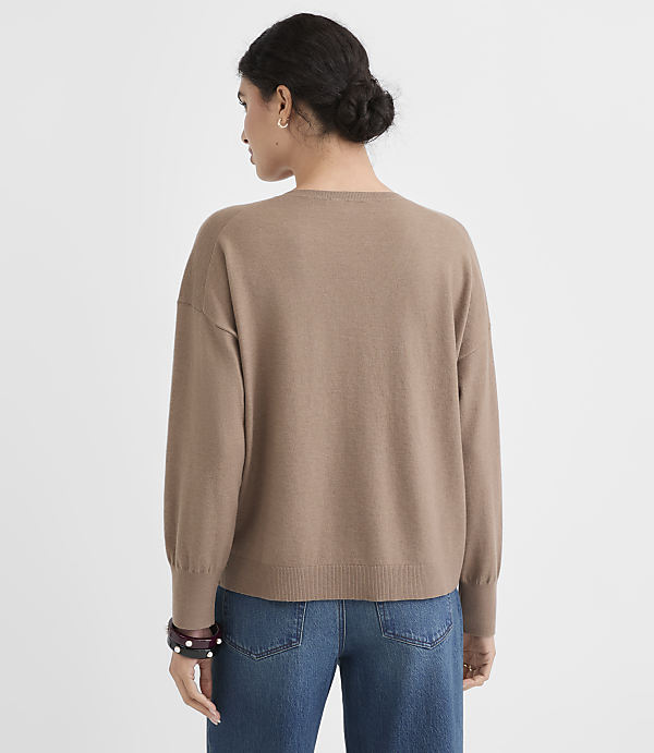 A woman in a tan sweater and blue jeans stands against a plain white background.