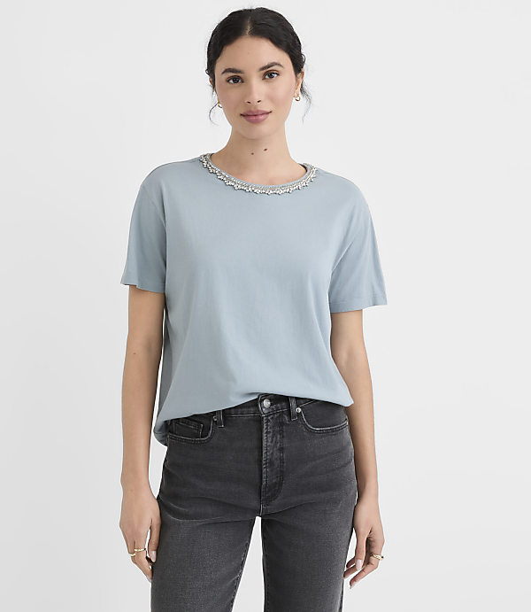 A woman in a light blue t-shirt with a decorative neckline and dark jeans stands against a plain white background.