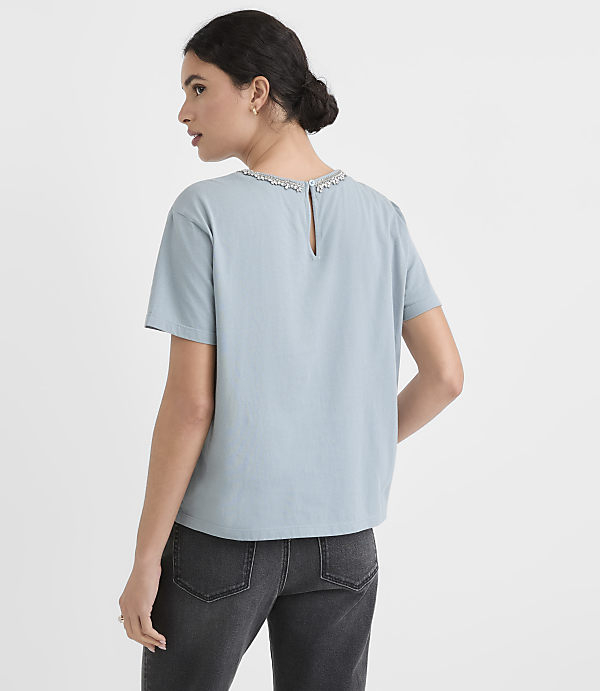 A woman in a light blue top and dark jeans stands against a plain white background.
