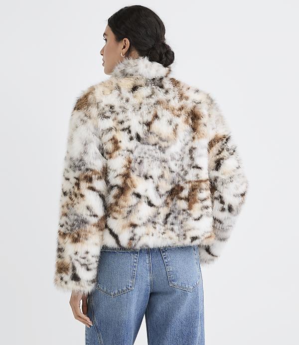 A woman in a spotted faux fur jacket and blue jeans stands against a plain white background.