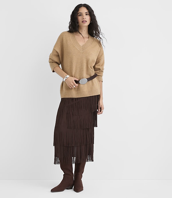 A woman in a tan v-neck sweater, brown fringed skirt, and brown boots stands against a plain white background.