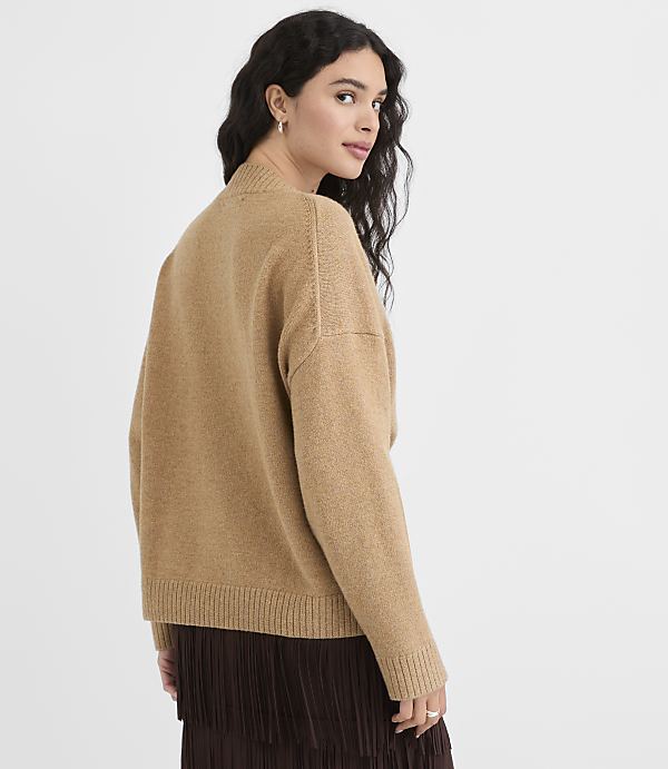A woman in a tan sweater and brown skirt stands against a plain, light background.