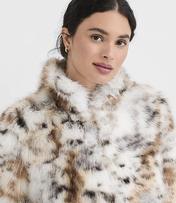 A woman with dark hair and light makeup wears a fluffy, spotted faux fur coat against a plain white background.