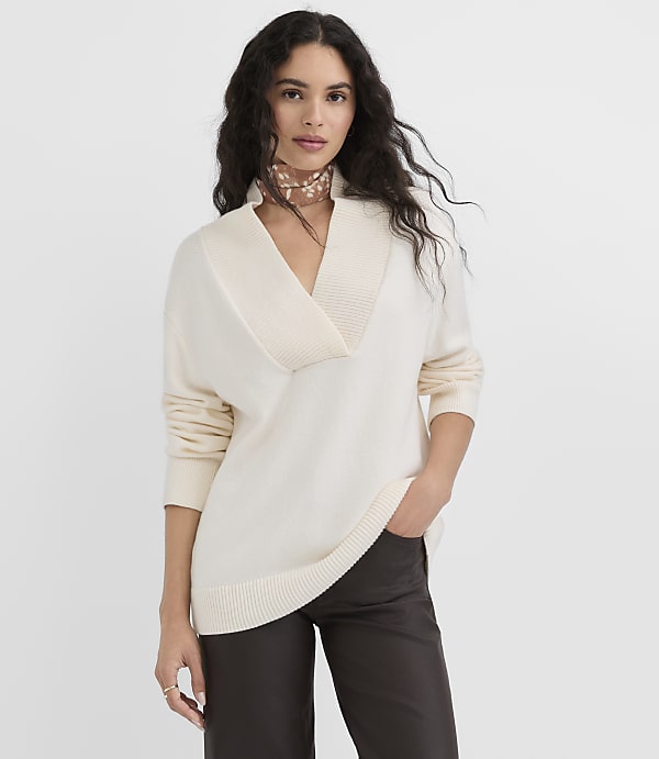A woman in a cream-colored sweater and dark pants stands against a plain gray background.