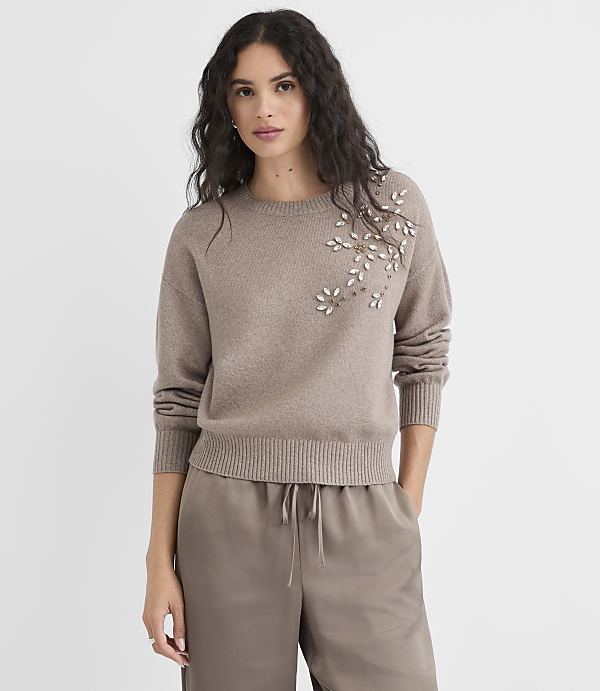 A woman in a taupe sweater with floral embellishments stands against a plain, light gray background.