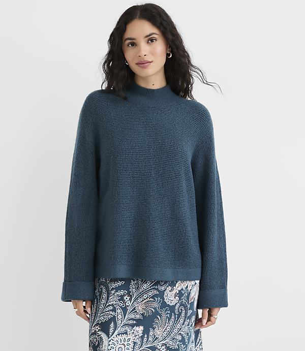 A woman in a teal sweater and patterned skirt stands against a plain white background.