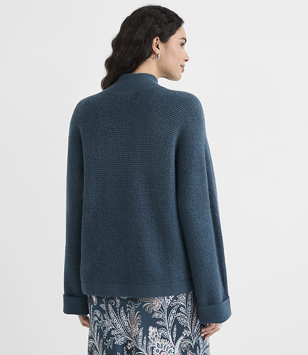A woman in a teal sweater and patterned skirt stands against a plain white background.