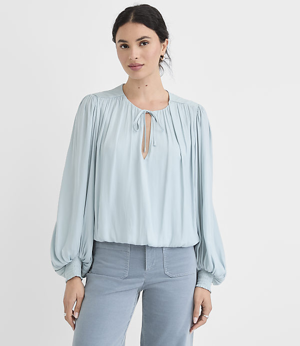 A woman in a light blue blouse and matching pants stands against a plain white background.