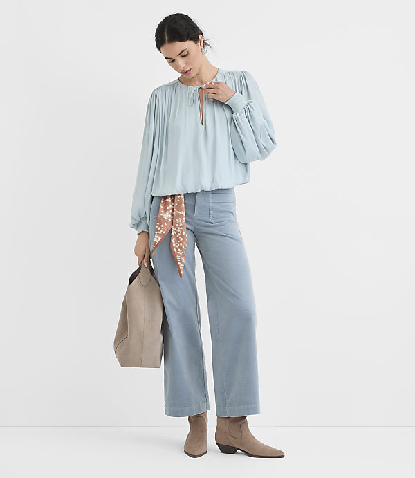 A woman in a light blue blouse and wide-leg pants stands against a plain white background, holding a tan tote bag.