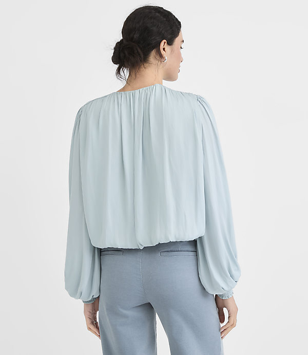 A woman in a light blue blouse and blue pants stands against a plain white background.