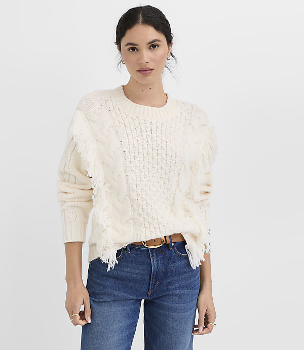 A woman in a cream cable-knit sweater with fringe detailing and blue jeans stands against a plain white background.
