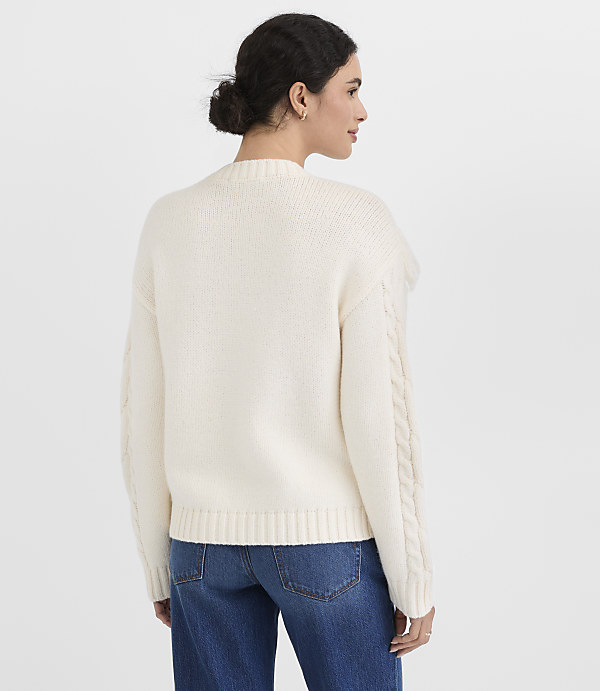 A woman in a cream cable-knit sweater and blue jeans stands against a plain white background.