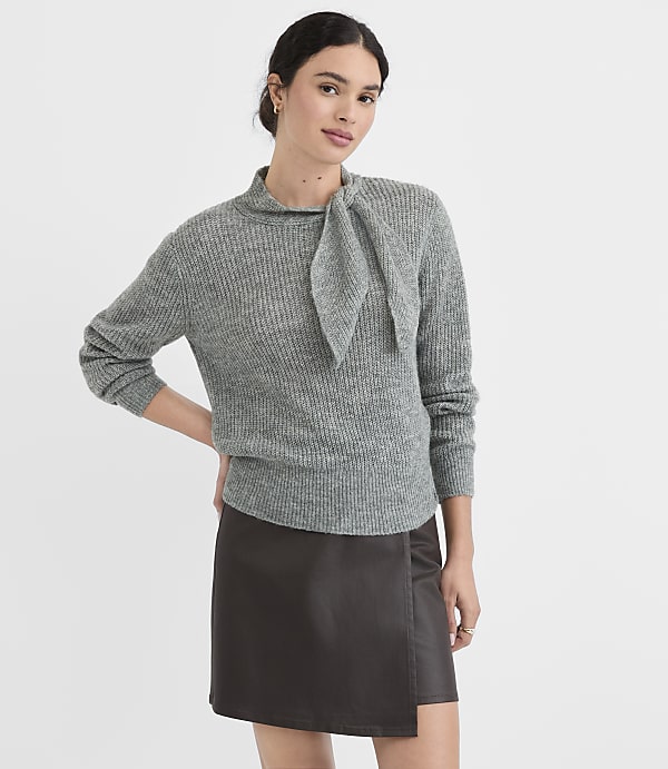 A woman in a grey sweater and dark skirt stands against a plain white background.