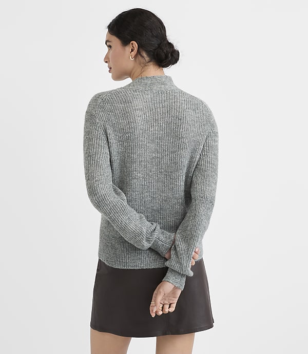 A woman in a grey ribbed sweater and dark skirt stands with her back to the viewer against a plain grey background.
