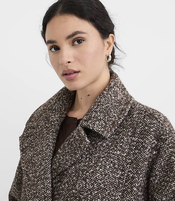 A woman in a textured brown coat with a white flecked pattern looks forward against a plain white background.