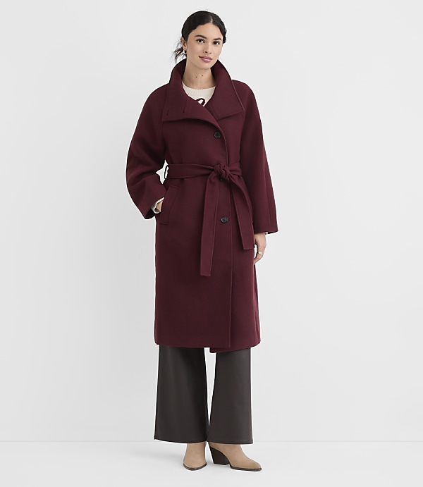 A woman in a burgundy coat and dark pants stands against a plain white background.