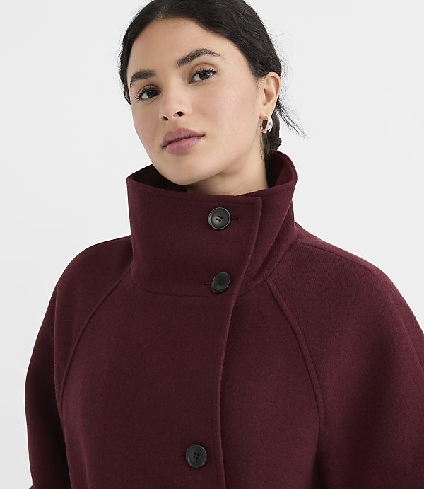 A woman in a burgundy coat with a high collar stands against a plain gray background.