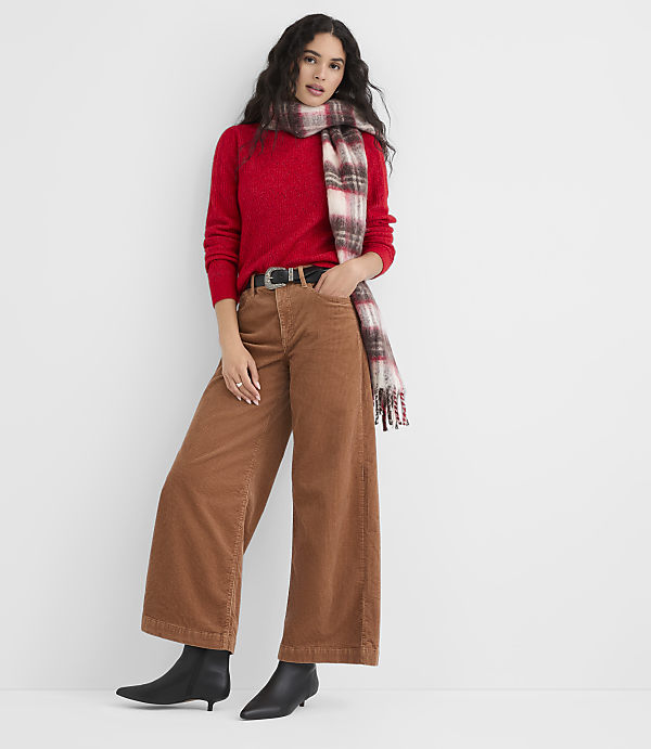 A woman in a red sweater, brown corduroy pants, and a plaid scarf stands against a plain white background.