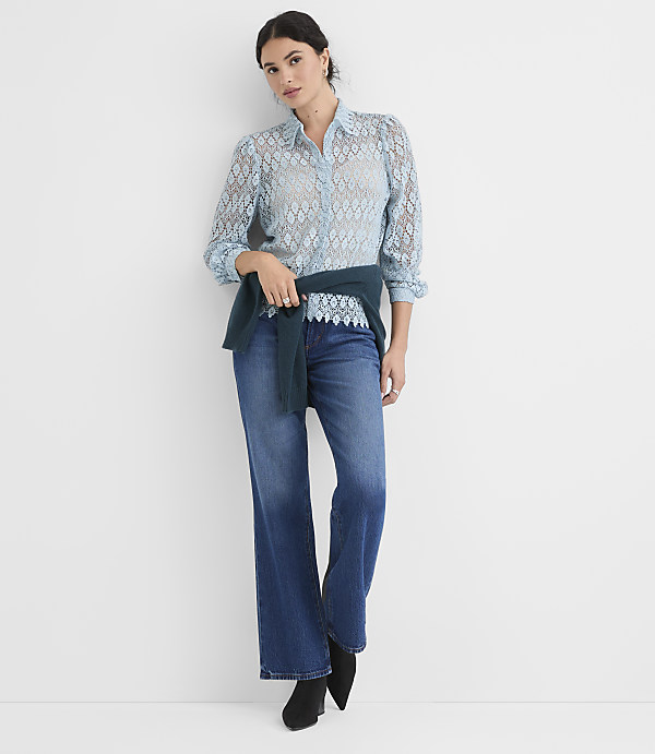 A woman in a light blue lace blouse and dark wash jeans stands against a plain white background.