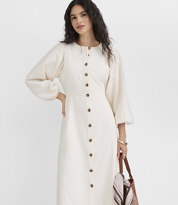 A woman in a cream-colored button-down dress with voluminous sleeves stands against a plain white background, holding a brown leather tote bag.