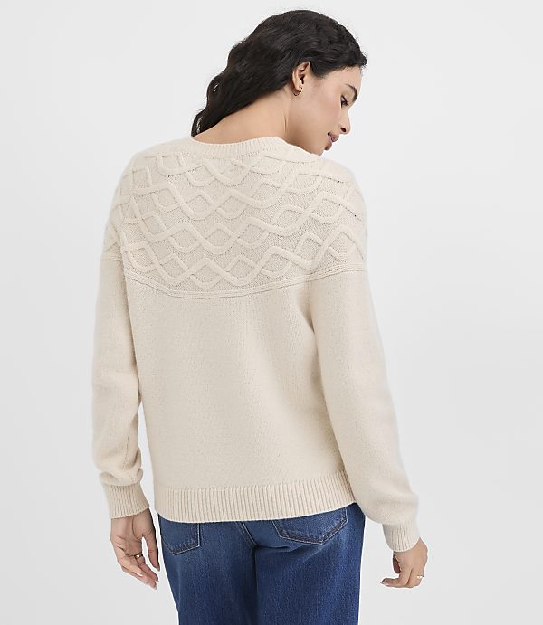 A woman in a cream-colored sweater and blue jeans stands against a plain white background.