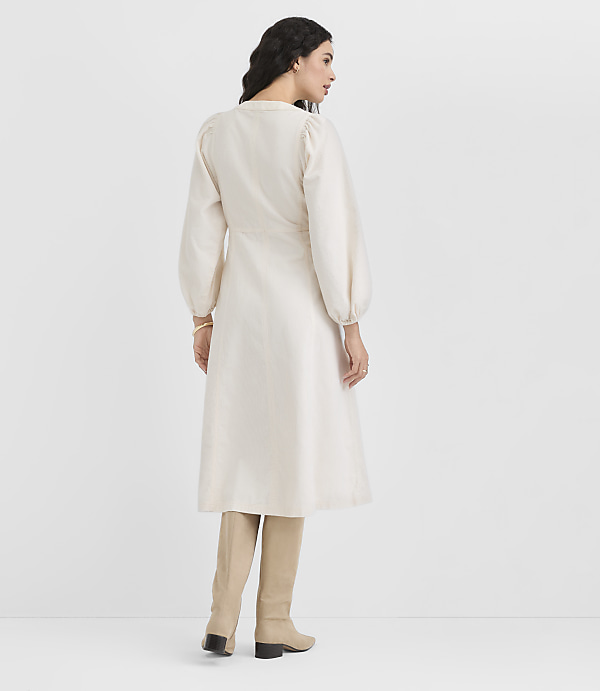 A woman in a cream-colored dress and beige boots stands against a plain white background.