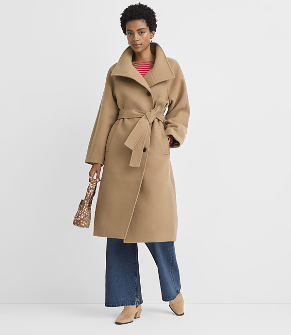 A woman in a tan coat and blue jeans stands against a plain white background, holding a patterned handbag.