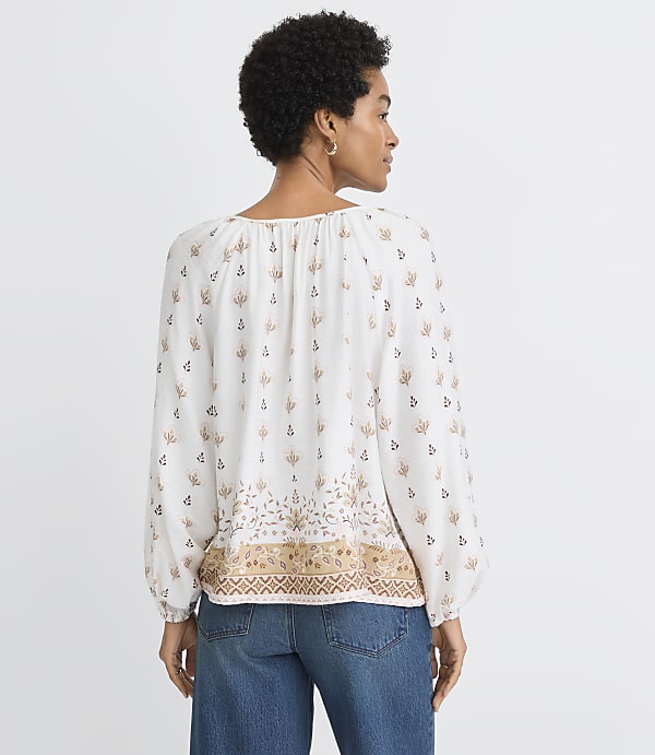 A woman in a patterned white blouse and blue jeans stands against a plain white background.