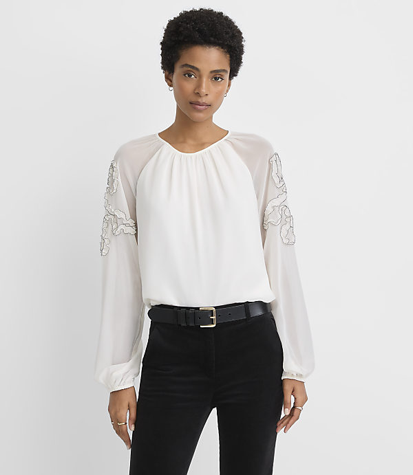 A woman in a white blouse and black pants stands against a plain white background.