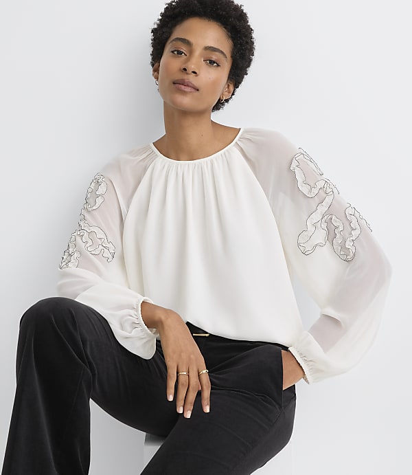A woman in a white blouse with sheer, embroidered sleeves and black pants sits against a plain white background.