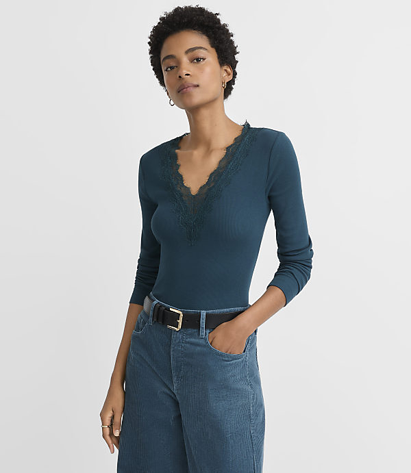 A woman in a teal ribbed long-sleeve shirt with lace trim and teal corduroy pants stands against a plain gray background.