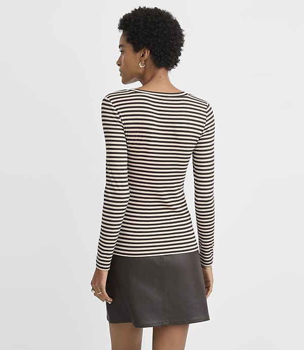 A woman in a striped long-sleeved shirt and a dark skirt stands against a plain, light gray background.