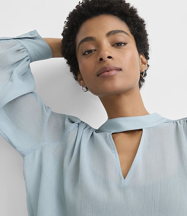 A woman with short, dark, curly hair and a light blue blouse with a keyhole neckline is shown against a plain, light gray background.