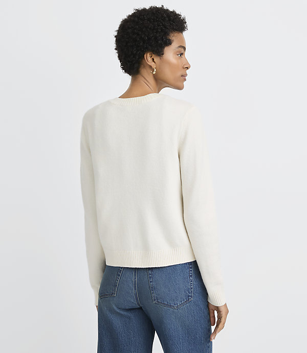 A woman in a cream sweater and blue jeans stands against a plain white background.