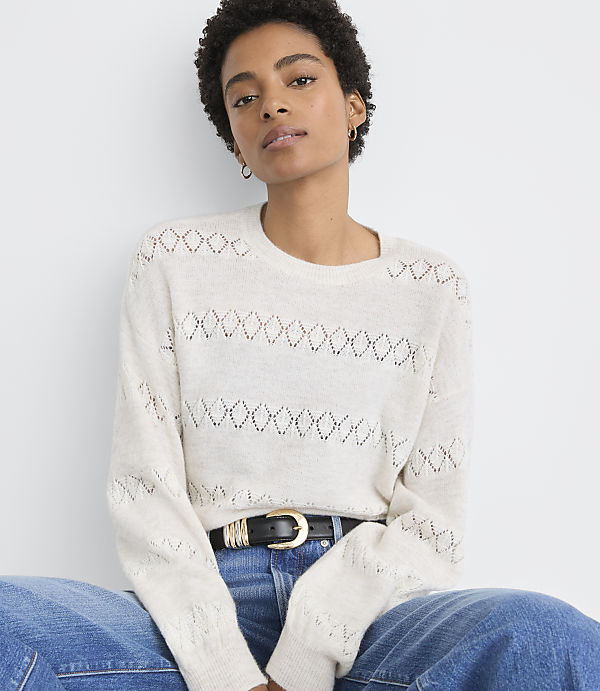 A woman in a cream-colored, open-knit sweater and blue jeans sits against a plain white background.