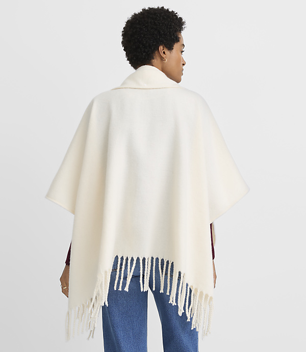 A woman wearing a cream-colored fringed poncho stands against a blurred background.