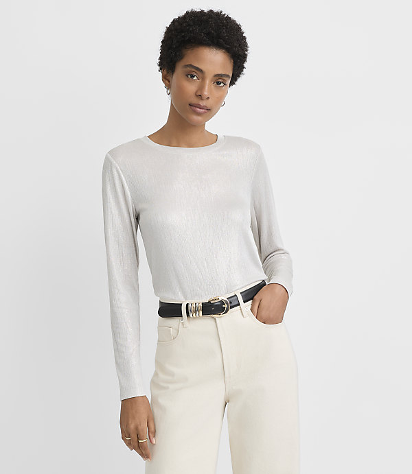 A woman in a shimmering silver long-sleeved shirt and cream pants with a black belt stands against a plain white background.