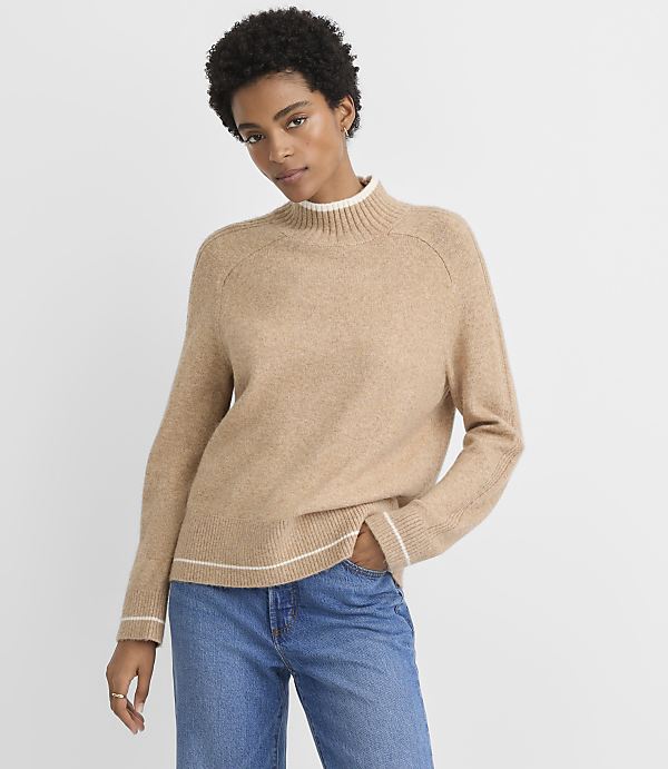 A woman in a tan mock-neck sweater and blue jeans stands against a plain gray background.