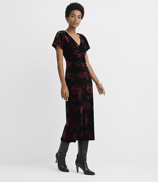 A woman in a black floral velvet dress and black boots stands against a plain white background.