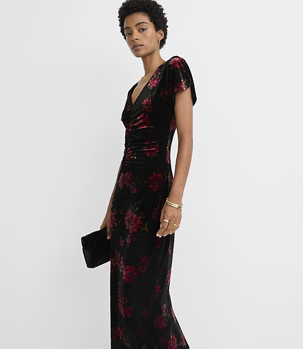 A woman in a black velvet floral dress holds a clutch in the foreground against a plain white background.