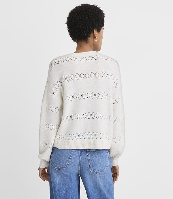 A woman in a cream-colored, open-knit sweater and blue jeans stands against a plain white background.