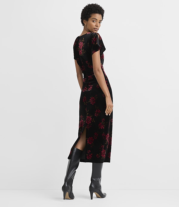 A woman in a black velvet dress with red floral print stands against a plain gray background.