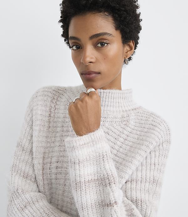 A woman with short, dark hair and a light-colored, textured sweater rests her chin on her hand against a plain, light background.