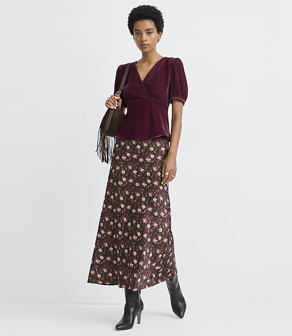 A woman in a burgundy velvet top and floral skirt stands against a plain white background.