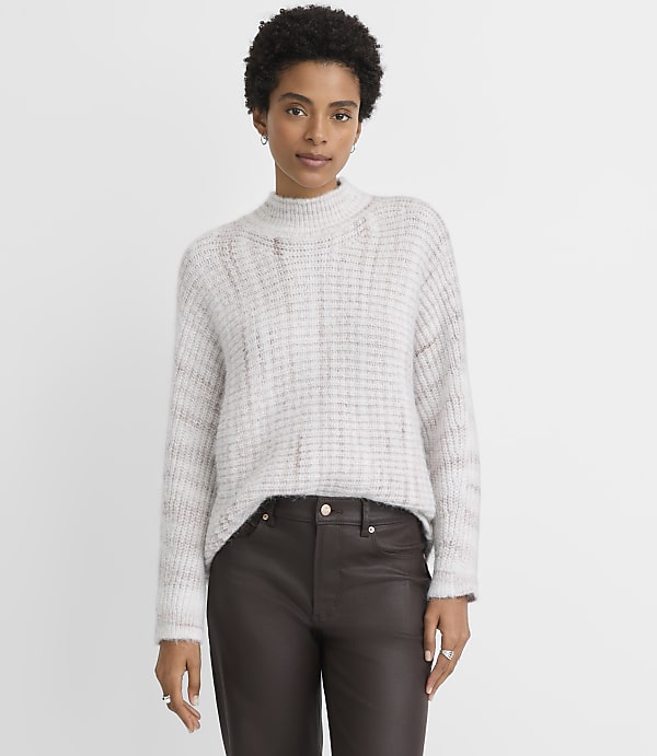A woman in a light-colored, textured sweater and dark pants stands against a plain, light gray background.