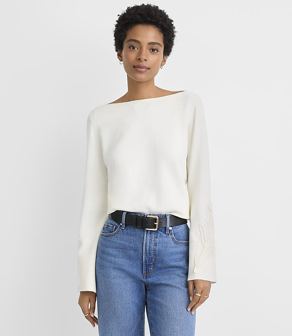 A woman in a white sweater and blue jeans stands against a plain white background.