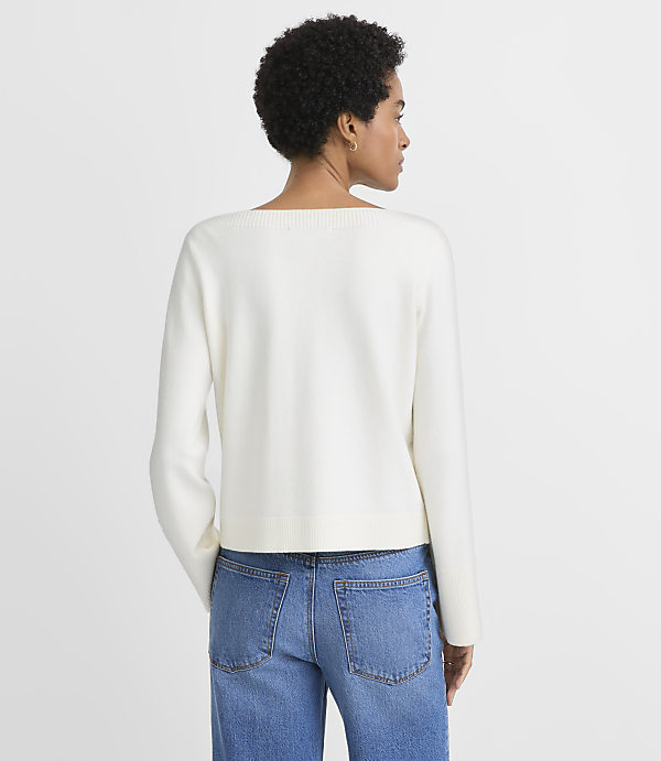 A woman in a white sweater and blue jeans stands against a plain white background.