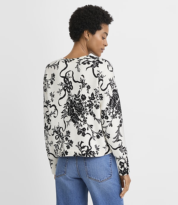 A woman wearing a black and white floral print sweater and blue jeans stands against a plain white background.