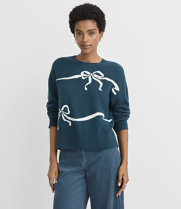 A woman in a teal sweater with white ribbon designs stands against a plain gray background.